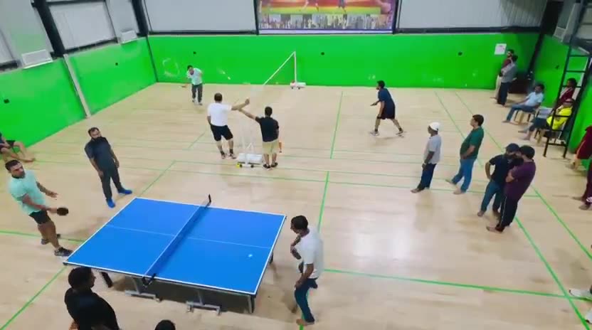 Close-up video of a Table Tennis rally in the foreground with Badminton doubles being played behind